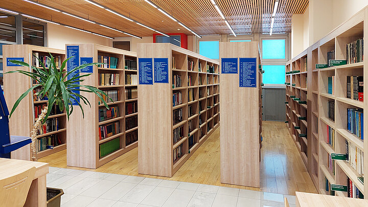 Zdjęcie przedstawia przestrzeń biblioteki, w której widać długie, jasne regały wypełnione książakmi.