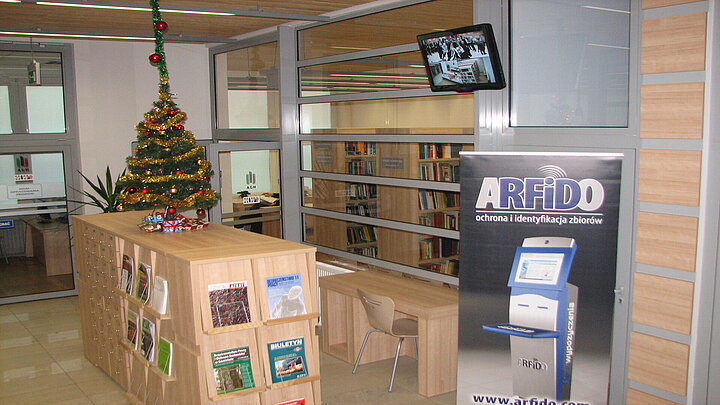 Hol biblioteczny, na środku szafka z wyeksponowanymi czasopismami, na szafce choinka świąteczna. Po lewej stronie od szafki stoi biurko z krzesłem przed szkalną ścianą, za którą widać regały z książkami.