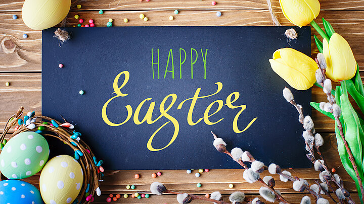 na stole leżą pisanki, bazie i tulipany a na środku kartka z napisem Happy Easter