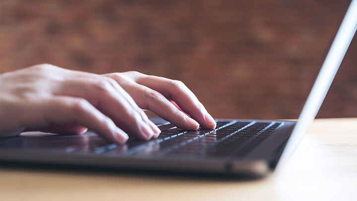 Na stole otwarty laptop z dłońmi na klawiaturze.