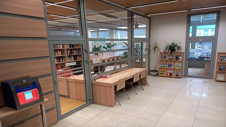Na zdjęciu hol biblioteki, z lewej strony wrzutnia na książki, dalej stół ustawiony przed szklaną ścianą, za którą znajduje się stanowisko bibliotekarza, wypożyczalnia i czytelnia.
