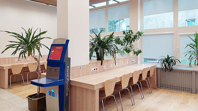 Pomieszczenie dla czytelników, widoczne stoły i krzesła oraz urządzenie do samodzielnego wypożyczania